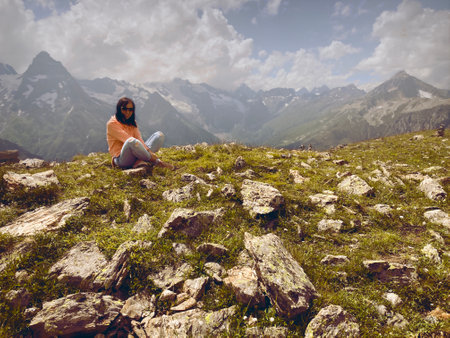 Young woman in sunglasses sitting on mountain top in sunny weather. Female tourist posing for camera on background of amazing mountain landscape in summertimeの写真素材