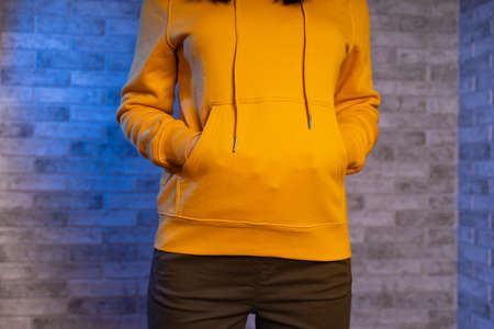 Close up of woman's hands of unrecognizable person in pockets of yellow hoodie on gray brick background. Front view on body part of young female in sweatshirtの写真素材