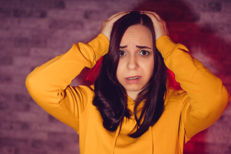 Portrait of unhappy young woman holding her head with hands. Close up of sad female in yellow hoodie clutching head from headacheの写真素材