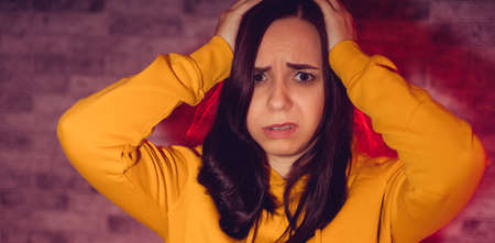 Portrait of unhappy young woman holding her head with hands. Close up of sad female in yellow hoodie clutching head from headacheの写真素材