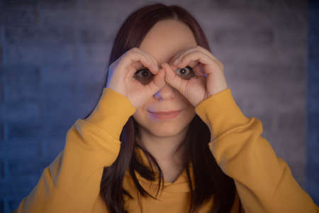 Portrait of young woman making binoculars out of her hands and smiling on gray brick background....