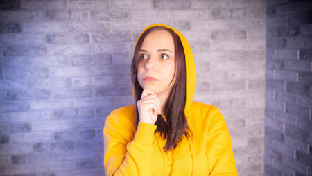 Portrait of thoughtful young woman in hood on gray brick background. Close up of cute brunette propping up her chin in reflections.の写真素材