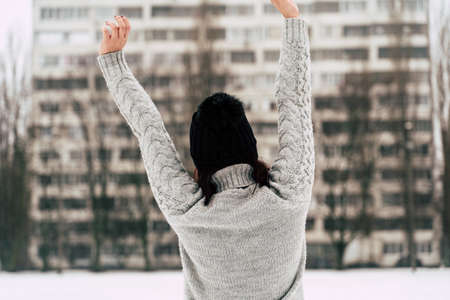 Rear view of young woman in gray knitted sweater and hat standing on street in winter season. Female looking at high-rise building and stretching her hands in cloudy weatherの写真素材