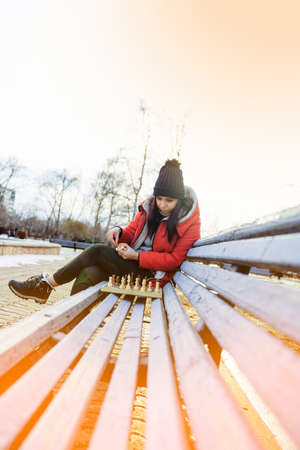 Young woman in winter clothes plays chess, sitting on bench in city park. Female in black cap with chess on wooden pew in cloudy weatherの写真素材