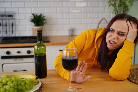 Unhappy young woman has headache from red wine. Adult female feels bad from alcohol while in kitchenの写真素材