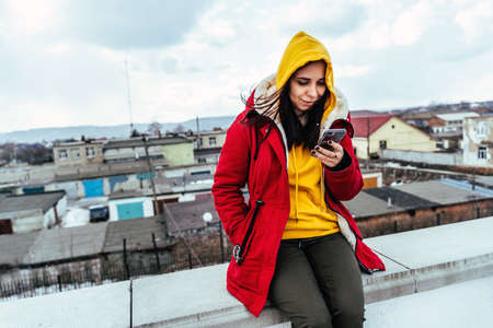 Portrait of young woman with mobile phone on roof of building. Brunette in yellow hoodie and red jacket browsing smartphone in cloudy weatherの写真素材
