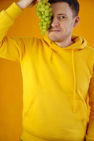 Portrait of young man with green grapes on yellow background. Close up of handsome guy in yellow hoodie closes eye of bunch of grapesの写真素材