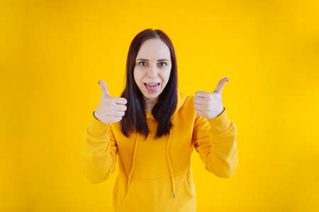 Portrait of young woman smiling and showing thumbs up on yellow background. Close up of happy brunette in yellow hoodie showing gesture of approvalの写真素材