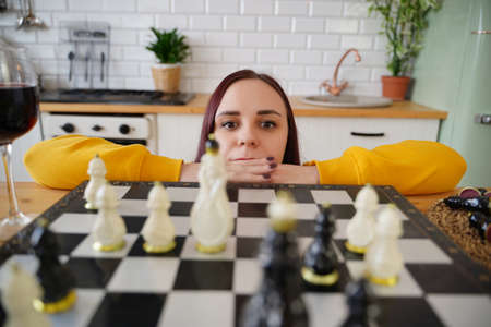 Young woman playing chess on kitchen table. Female plays in logical board game with herself in kitchenの写真素材