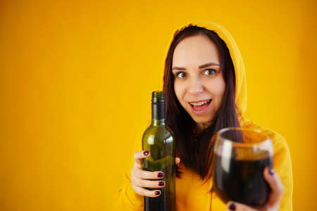 Relaxed young woman poses with bottle and glass of red wine on yellow background. Adult happy brunette holds alcohol in handsの写真素材