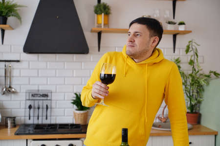 Relaxed young man poses with glass of red wine, standing at kitchen table. Adult guy resting with alcohol in kitchenの写真素材