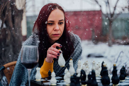 Young woman playing chess in yard. Female wrapped in gray plaid sitting on street playing in board game in winter seasonの写真素材