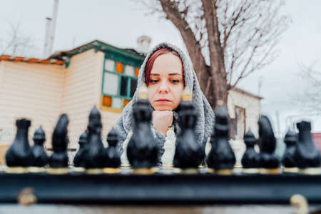 Young woman playing chess in yard. Female wrapped in gray plaid sitting on street playing in board game in winter seasonの写真素材
