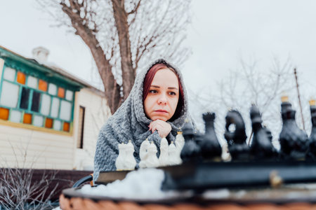 Young woman playing chess in yard. Female wrapped in gray plaid sitting on street playing in board game in winter seasonの写真素材