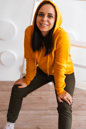 Portrait of young woman posing on white background. Pretty brunette in yellow hoodie near stepladder...