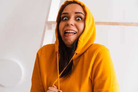 Portrait of young woman posing on white background. Pretty brunette in yellow hoodie near stepladder...