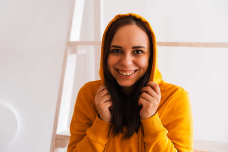 Portrait of young woman posing on white background. Pretty brunette in yellow hoodie near stepladder on background of white wallの写真素材