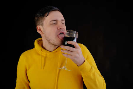 Portrait of young man licking glass with red wine on black background. Passionate guy in yellow hoodie with wine glass on dark backgroundの写真素材