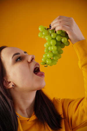 Portrait of young woman with bunch of grapes on yellow background. Close up of female opens mouth, going to eat green grapesの写真素材