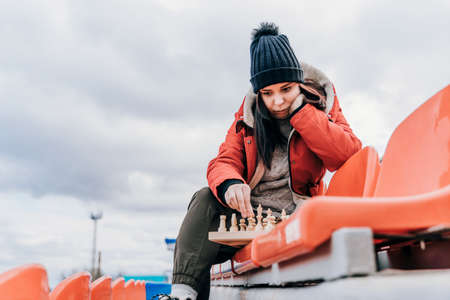 Young woman in winter clothes plays chess, sitting on stadium bleachers alone. Female in black cap with chess on sports stadium in cloudy weatherの写真素材