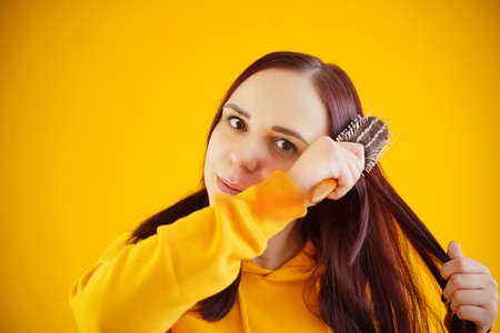 Portrait of young woman combing her hair on yellow background. Brunette in yellow hoodie preens, while looking at cameraの写真素材