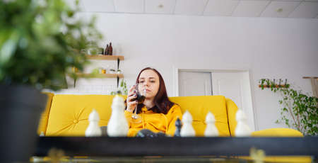 Young woman playing chess and drinking red wine in room. Adult female with alcohol plays in logical board game with herself, sitting on sofaの写真素材