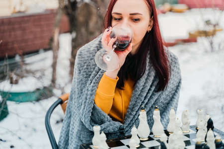 Young woman playing chess and drinking red wine in yard. Female wrapped in gray plaid with alcohol...