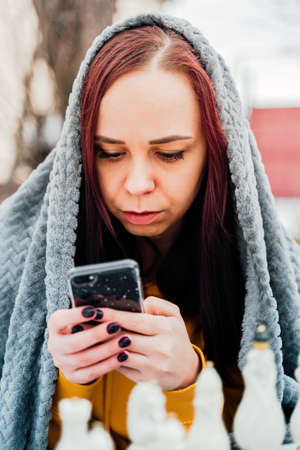 Young woman playing chess and browsing smartphone in yard. Female wrapped in gray plaid with mobile phone sitting on street playing in board game in winter seasonの写真素材