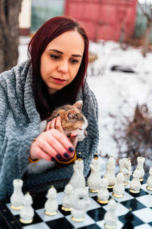 Young woman sitting with cat and playing chess in yard. Female wrapped in gray plaid sitting on street with pet and playing in board game in winter seasonの写真素材