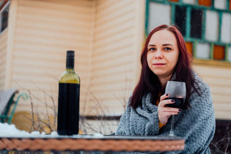 Young woman poses, sitting on street in yard. Female wrapped in gray plaid with alcohol while picnic in winter seasonの写真素材