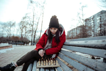 Young woman in winter clothes plays chess, sitting on bench in city park. Female in black cap with...