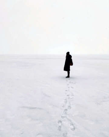 Unrecognizable woman in black clothes standing on snowy lake in cloudy weather. Silhouette of female walking on winter lake in cold weatherの写真素材