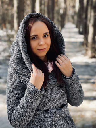 Portrait of young beautiful woman in gray coat on walk in forest. Close up of pretty brunette in hood posing on sunny day in early springの写真素材