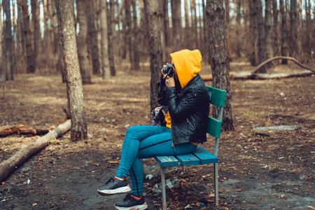 Young woman in casual clothes sitting on bench and photographing on old photo camera in forest....