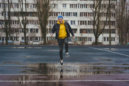 Young man runs through puddle on sports stadium on background of high-rise building. Happy guy with blue hair having fun on city street in springtimeの写真素材