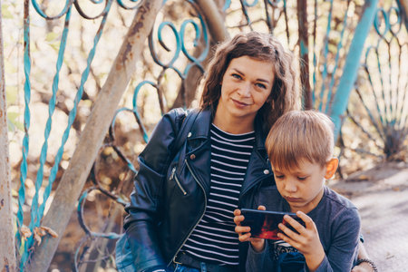 A focused boy is playing a game on a smartphone in the park. A serious focused school age boy in casual clothes is sitting on a bench and together with his mother and playing a game on a mobile phoneの写真素材