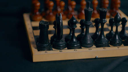 Close up of wooden black chess pieces on chessboard. Unknown female hand makes first move of white...