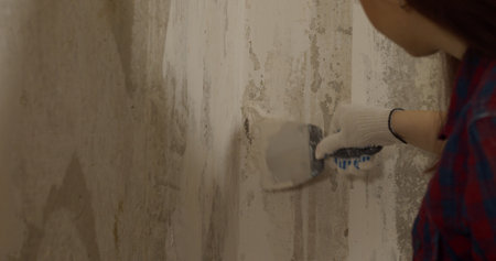 Close up of young woman with gloves applying putty on wall in room. Female plastering wall with...