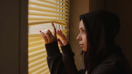 Close up of young woman watching through window blinds at home. Portrait of curious female observing...