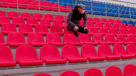 Young man in medical mask sitting on stadium bleachers alone. Sad adult male protecting yourself from diseases at sports stadium. Concept of restrictions on public events and competitionsの写真素材