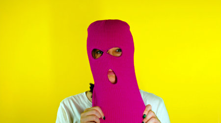 Close up young woman puts on pink balaclava on yellow background. Secretive female puts on mask, looking at cameraの写真素材