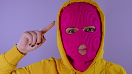 Close up of young woman in pink balaclava and yellow hood twirling finger at her temple on purple background. Surprised female in mask twists index finger at temple, looking at cameraの写真素材