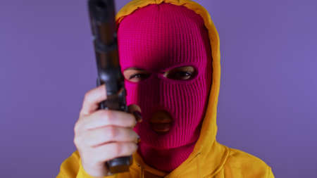 Unrecognizable woman in pink balaclava with gun on purple background. Dangerous female in mask takes aim at camera, shoots and laughs. Slow motion.の写真素材