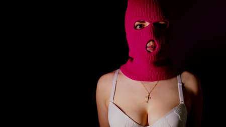Unrecognizable woman in pink balaclava and white bra stands in dark. Unknown female in mask and lingerie highlighted in dark roomの写真素材