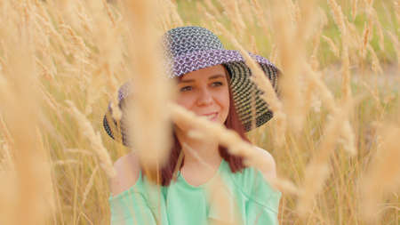 Young woman in dress and hat sitting in wheat field. Pretty lady hides, sitting in ears of wheat.の写真素材