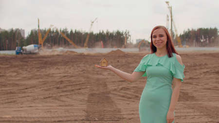 Young woman with small wooden house in hand stands on construction site. Concept of purchasing new...