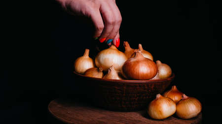 Pile of whole bulbs of raw onion in ceramic bowl on table. Woman's hand takes bulb of onion from bowlの写真素材