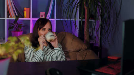 Young woman drinks coffee, sitting in armchair before computer in office. Employee enjoying hot...