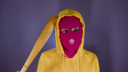 Young woman in pink balaclava with baseball bat. Hooligan in mask scratching head with bat and looking at camera.の写真素材