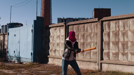 Young woman in pink balaclava with baseball bat in hands. Hooligan in mask beats with bat, looking at camera.の写真素材